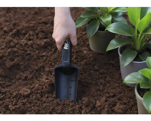 Eine Hand hält eine Pflanzschaufel in der Erde, neben eingetopften Pflanzen.