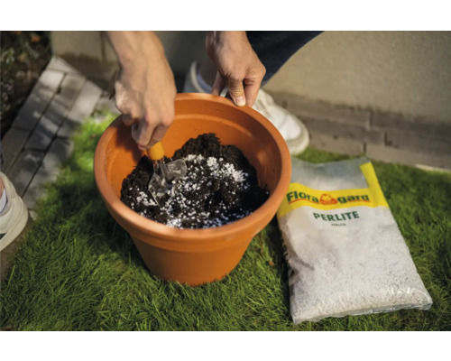 Eine Person mischt Blumenerde mit Perlit in einem Pflanzgefäß. Eine Packung Flora Gard Perlite liegt daneben.