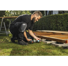 Ein Mann baut eine Holzterrasse mit einem Akku-Bohrschrauber und Knieschützern im Garten.