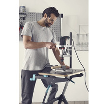 Mann bohrt mit einer Bohrmaschine und einem Bohrständer ein Werkstück auf einer Werkbank.