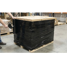 Eine Palette mit Kartons, verpackt in schwarzer Stretchfolie, wird von einer Person mit einem Handabroller gesichert.