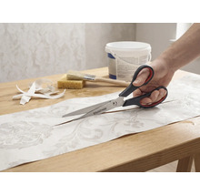 Eine Person schneidet ein Stück gemusterte Tapete mit einer Schere der Marke Rautner auf einem Tisch