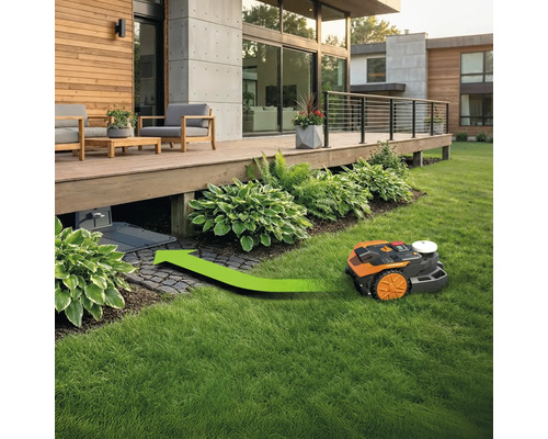 Ein Rasenmähroboter auf einem Rasen mit einer grünen Spur, die zu einer Ladestation unter einer Terrasse führt.