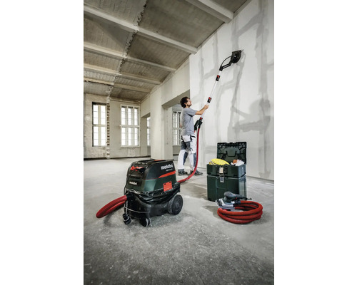 Mann schleift Wand mit einem Langhalsschleifer, Staubsauger und Werkzeugkoffer im Raum