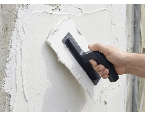 Eine Person trägt mit einer Glättkelle Putz auf eine Wand auf.