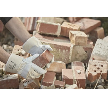 Eine Person mit Arbeitshandschuhen hält einen Ziegelstein vor einem Haufen Ziegelsteine.