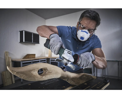 Anwender mit Atemschutzmaske, Schutzbrille und Handschuhen bearbeitet Holz mit einem Winkelschleifer