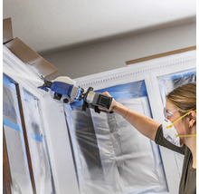 Frau streicht Fensterrahmen mit Farbsprühgerät, Fenster sind mit Folie abgedeckt