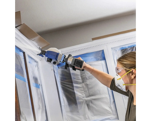 Frau streicht Fensterrahmen mit Farbsprühgerät, Fenster sind mit Folie abgedeckt