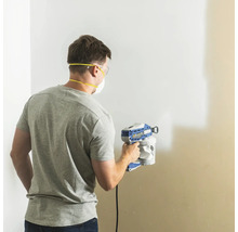 Mann streicht Wand mit Farbsprühgerät, ausgestattet mit Schutzbrille und Staubmaske