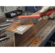 Eine Hand mit Arbeitshandschuh reinigt ein rostiges Stahlprofil mit einer Drahtbürste.