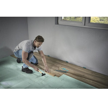 Ein Mann verlegt Laminatboden mit Hammer und Schlagholz auf Trittschalldämmung in einem Raum.
