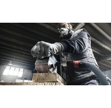 Ein Mann schleift mit einer Schleifmaschine ein Stück Holz. Er trägt eine Schutzmaske und Schutzbrille.