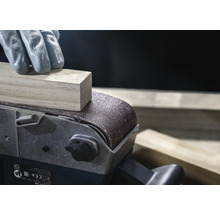 Nahaufnahme eines Bandschleifers beim Schleifen eines Holzstücks, bedient von einer Person mit Handschuhen.