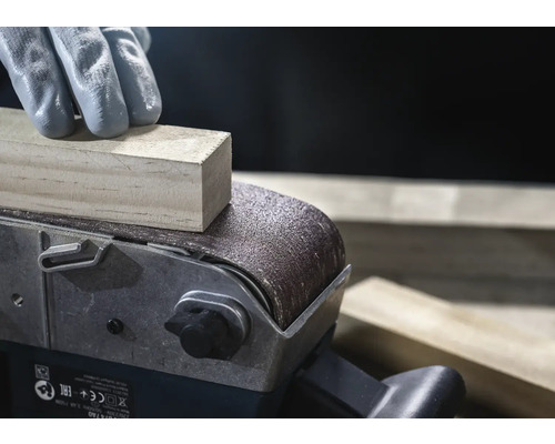 Nahaufnahme eines Bandschleifers beim Schleifen eines Holzstücks, bedient von einer Person mit Handschuhen.
