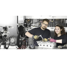 Zwei Personen reparieren einen Motor in einer Werkstatt mit Motorrädern und Werkzeugen.