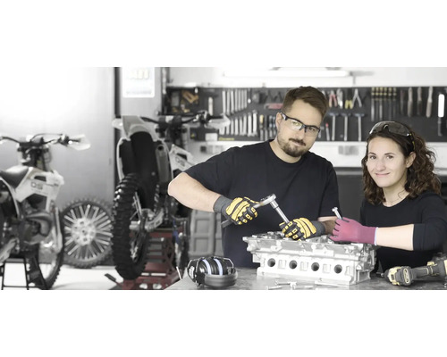 Zwei Personen reparieren einen Motor in einer Werkstatt mit Motorrädern und Werkzeugen.