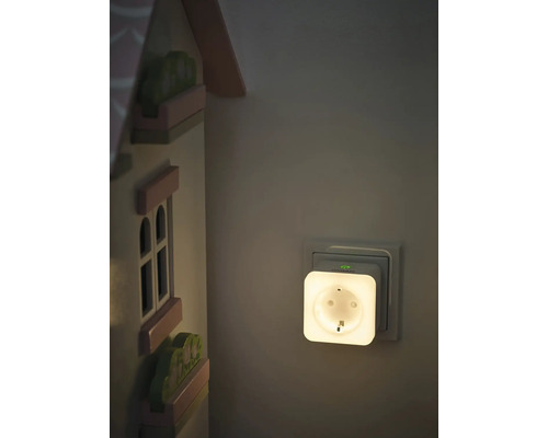 OSRAM Smart Plus Plug Nachtlicht in einer Wandsteckdose neben einem hölzernen Spielzeughaus in einem sanft beleuchteten Raum.
