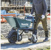 Eine Person schiebt eine mit Erde gefüllte Makita Akku-Scheibtruhe auf einer hellen Baustelle. Blaues Gerät mit drei Rädern und Makita Logo.
