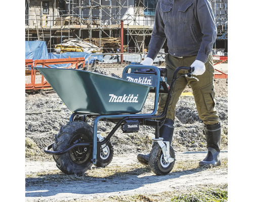 Eine Person schiebt eine mit Erde gefüllte Makita Akku-Scheibtruhe auf einer hellen Baustelle. Blaues Gerät mit drei Rädern und Makita Logo.