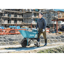 Bauarbeiter schiebt eine Makita Akku-Schubkarre auf einer Baustelle mit Gerüsten im Hintergrund.