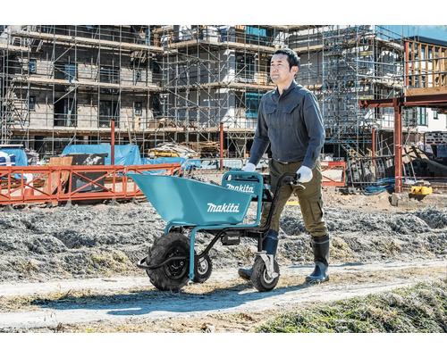 Bauarbeiter schiebt eine Makita Akku-Schubkarre auf einer Baustelle mit Gerüsten im Hintergrund.