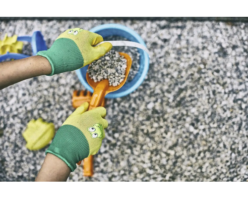Hände mit Gartenhandschuhen befüllen einen Eimer mit einer Schaufel und Steinen, umgeben von Spielzeug für den Sandkasten.