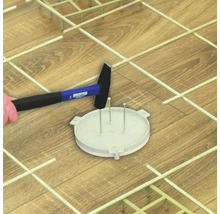 Ein Hammer schlägt auf eine runde Kunststoff-Grundplatte mit Stiften auf einem Holzboden inmitten einer Gitterstruktur zur Bodenverlegung.