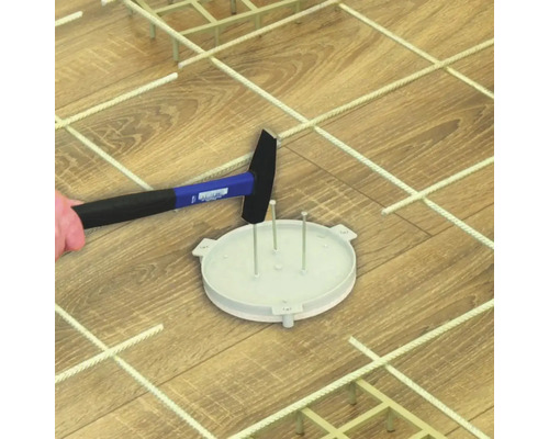 Ein Hammer schlägt auf eine runde Kunststoff-Grundplatte mit Stiften auf einem Holzboden inmitten einer Gitterstruktur zur Bodenverlegung.