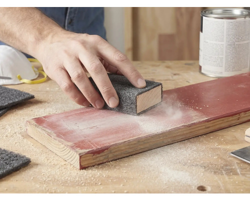 Hand schleift ein lackiertes Holzbrett mit einem Schleifvlies auf einem Schleifklotz. Schleifstaub ist auf dem Holz sichtbar.