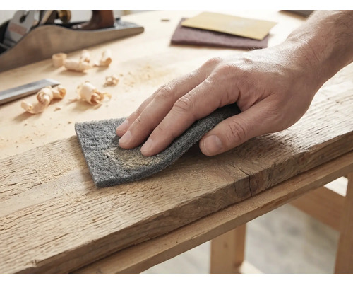 Hand bei der Bearbeitung eines Holzbretts mit Schleifvlies auf einer hellen Werkbank mit Hobel und Holzspänen.