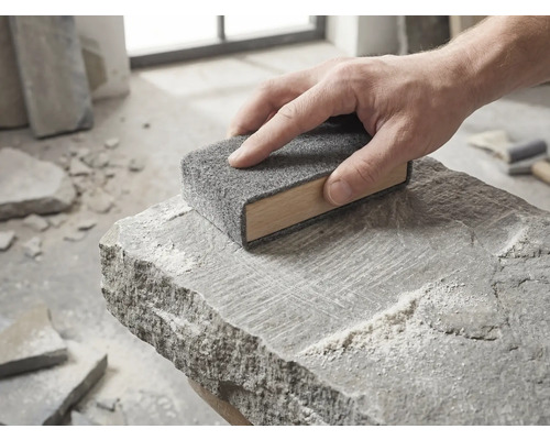 Eine Hand führt einen hölzernen Schleifklotz mit grauem Schleifvlies über die Oberfläche eines rauen Natursteins.