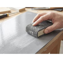 Eine Hand schleift eine lackierte Holzoberfläche mit einem Schleifvlies, das um einen Schleifklotz gewickelt ist.