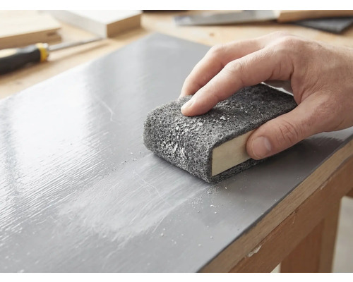 Eine Hand schleift eine lackierte Holzoberfläche mit einem Schleifvlies, das um einen Schleifklotz gewickelt ist.