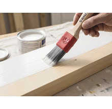 Ein Pinsel mit Holzgriff und roter Manschette trägt weiße Farbe auf einen Holzbalken auf. Im Hintergrund steht eine geöffnete Farbdose.