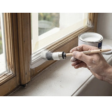 Hand streicht einen Holzfensterrahmen weiß mit einem Rautner Ringpinsel, neben einer geöffneten Farbdose.