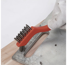 Hand mit Arbeitshandschuh reinigt eine Metalloberfläche mit einer kleinen Drahtbürste mit orangem Griff.