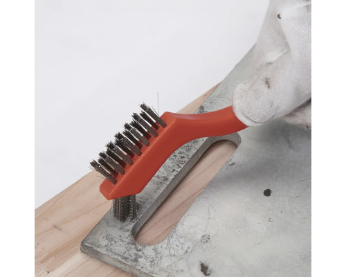 Hand mit Arbeitshandschuh reinigt eine Metalloberfläche mit einer kleinen Drahtbürste mit orangem Griff.