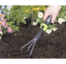 Eine Hand führt eine for_q Kleingartenharke durch dunkle Erde in einem Beet mit bunten Blumen.