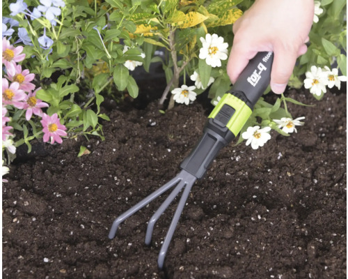 Eine Hand führt eine for_q Kleingartenharke durch dunkle Erde in einem Beet mit bunten Blumen.