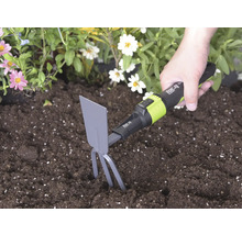 Hand führt einen for_q Kleinhäcker mit Zinken und Blatt in dunkler Erde neben Blumen zum Lockern des Bodens.