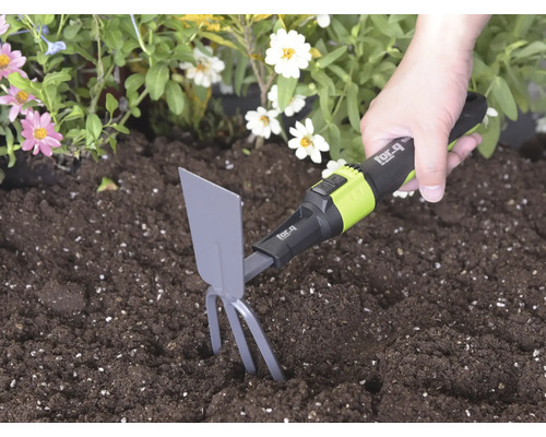 Hand führt einen for_q Kleinhäcker mit Zinken und Blatt in dunkler Erde neben Blumen zum Lockern des Bodens.