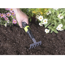 Handrechen mit schwarzem und grünem Griff und for_q Logo wird in dunkler Blumenerde zwischen rosa und weißen Blüten verwendet.