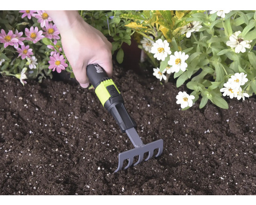 Handrechen mit schwarzem und grünem Griff und for_q Logo wird in dunkler Blumenerde zwischen rosa und weißen Blüten verwendet.