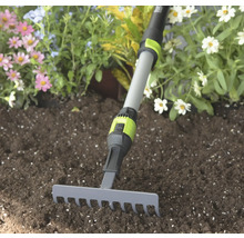 Kleiner Gartenrechen mit grauem Metallkopf und verstellbarem Teleskopstiel in dunkler Erde vor Blumen.