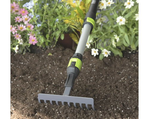 Kleiner Gartenrechen mit grauem Metallkopf und verstellbarem Teleskopstiel in dunkler Erde vor Blumen.