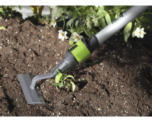 Garten-Pendelhacke mit grauer Klinge und grünem Anschlussstück bei der Unkrautentfernung in dunkler Erde neben einer Pflanze.