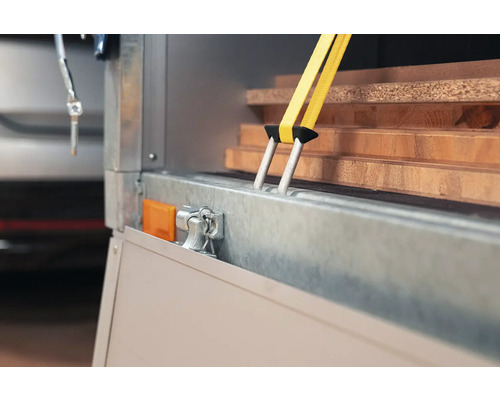 Gelber Spanngurt fixiert Holzbretter auf einem Anhänger zur Ladungssicherung mit Doppelhaken aus Metall.