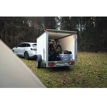 Weißer Humbaur Kofferanhänger an einem Auto, beladen mit einem Mountainbike und Ausrüstung in einem Waldgebiet.