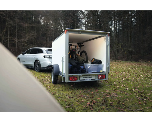 Weißer Humbaur Kofferanhänger an einem Auto, beladen mit einem Mountainbike und Ausrüstung in einem Waldgebiet.